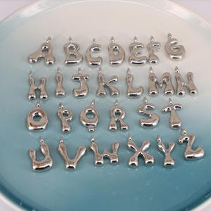May include: A selection of silver alphabet charms, each with a rounded, bubble-like design. The letters A to Z are displayed, each with a small loop at the top for attaching to a chain or bracelet. The charms are arranged on a light blue plate.