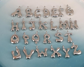 May include: A collection of silver-colored alphabet charms, each with a rounded, bubble-like design. The letters A through Z are displayed, each with a small loop at the top for attaching to a chain or bracelet. The charms are arranged on a light blue plate.