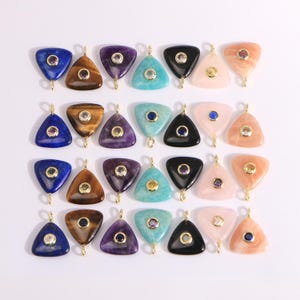May include: A collection of triangular gemstone pendants in various colors, including blue, brown, purple, turquoise, black, and pink. Each pendant has a gold-colored bail and a small, central gemstone. The pendants are arranged on a white surface.