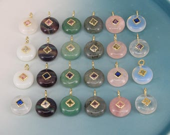 Puede incluir: Una colección de colgantes de piedras preciosas en varios colores, incluyendo blanco, morado, verde, gris, rosa y azul. Cada colgante presenta una argolla dorada y una pequeña gema cuadrada en el centro. Los colgantes están dispuestos sobre un plato azul claro.