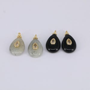 May include: Four teardrop-shaped pendants. Two are light gray with a speckled pattern, and two are black. Each pendant has a small, oval-shaped, light-colored stone set in a gold-colored bezel. A gold-colored loop is at the top of each pendant.