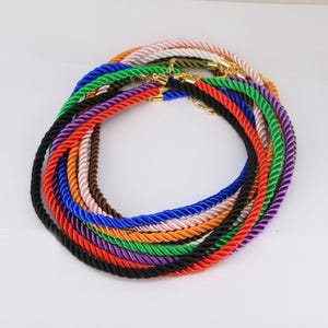 May include: A collection of colorful twisted ropes in various shades, including black, red, green, and blue. Each rope has gold-colored metal clasps. The ropes are arranged in a circular pattern against a white background.