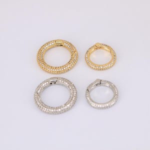 May include: Four round, hinged clasps in gold and silver tones, encrusted with small, sparkling stones. Two are gold, and two are silver. The clasps vary in size, with two larger and two smaller. The clasps are on a white background.
