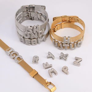 May include: Silver and gold-toned mesh bracelets with sparkling, letter-shaped charms. Individual letter charms are also displayed. The bracelets have a clasp closure. The letters are encrusted with small, clear stones.
