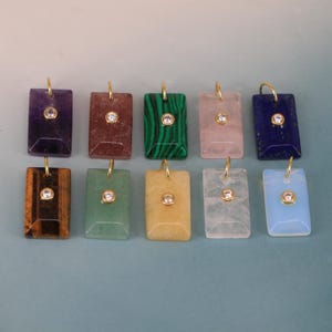 May include: Ten rectangular gemstone pendants arranged in two rows. Each pendant features a polished stone in various colors including purple, brown, green, pink, blue, tiger eye, and clear, each accented with a central round clear stone and gold bail.