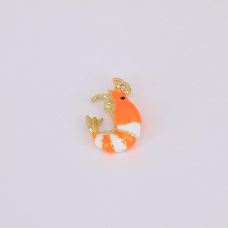 May include: A gold-colored shrimp charm with orange and white stripes. The shrimp has a small black eye and a gold-colored hook at the top for attaching to a necklace or bracelet. The charm is a cute and whimsical accessory.