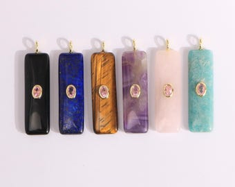 Puede incluir: Seis colgantes rectangulares de piedras preciosas en negro, azul, marrón, morado, rosa y turquesa. Cada colgante tiene una argolla dorada y una pequeña gema rosa en el centro. Los colgantes están dispuestos en fila sobre un fondo blanco.