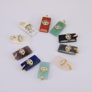 May include: A collection of rectangular pendants in various colors, including red, green, and blue, each adorned with a gold-colored bail and a decorative eye charm. The charms feature a blue eye design with surrounding embellishments.