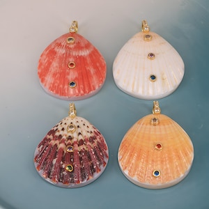 Könnte beinhalten: Vier Muschelanhänger mit goldfarbenen Ösen, jeweils mit drei eingelassenen runden Edelsteinen in verschiedenen Farben. Die Muscheln umfassen eine rote, eine weiße, eine dunkelbraun-weiß gestreifte und eine hellorangefarbene, alle poliert.
