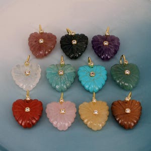 May include: A collection of eleven heart-shaped gemstone pendants, each featuring a carved radial pattern and a central round clear stone set in a gold-toned metal bail. The pendants display various colours including red, green, purple, white, blue, and pink.