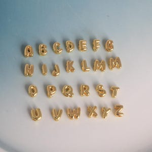 May include: A collection of gold-colored alphabet charms, each letter in a rounded, bubble-like font. The letters are arranged in rows, showcasing the entire alphabet from A to Z. The charms have a shiny, metallic finish.