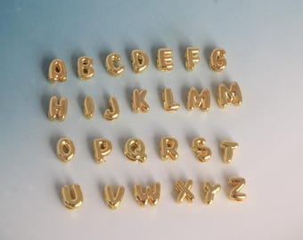 May include: A collection of gold-colored alphabet charms, each letter in a rounded, bubble-like font. The letters are arranged in rows, showcasing the entire alphabet from A to Z. The charms have a shiny, metallic finish.