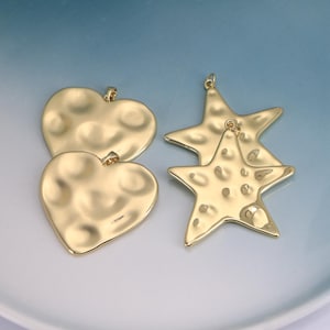 May include: Gold-colored metal pendants in heart and star shapes. The heart pendants have a hammered texture, and the star pendants have a similar hammered texture. The pendants are displayed on a white and blue surface.