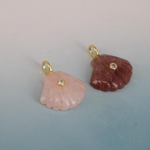 May include: Two seashell pendants. One is light pink, the other is reddish-brown. Each has a small, clear gem in the center and a gold-colored loop at the top for a chain. The pendants are on a light blue and white background.