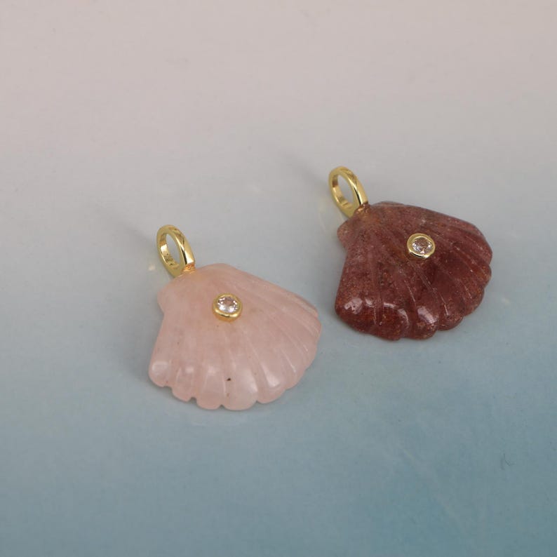 May include: Two seashell pendants. One is light pink, the other is reddish-brown. Each has a small, clear gem in the center and a gold-colored loop at the top for a chain. The pendants are on a light blue and white background.