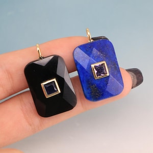 Puede incluir: Dos colgantes rectangulares con aros dorados. Uno es negro con un centro cuadrado azul oscuro, el otro es azul con un centro cuadrado púrpura. Ambos tienen superficies facetadas y se muestran sobre un fondo azul claro.