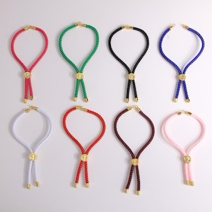 May include: A collection of eight bolo necklaces in various colors, including red, green, black, blue, white, and pink. Each necklace features a rope design with gold-colored metal accents and a central decorative element.