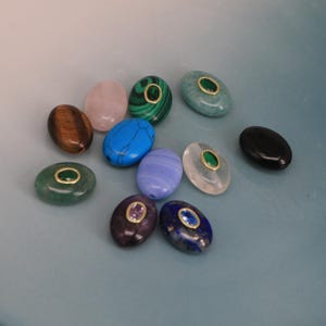 Puede incluir: Una colecci&oacute;n de cuentas de piedras preciosas de forma ovalada en varios colores, incluyendo verde, azul, rosa y marr&oacute;n. Algunas cuentas tienen un anillo dorado alrededor de una piedra m&aacute;s peque&ntilde;a. Las cuentas est&aacute;n dispuestas sobre una superficie azul claro.