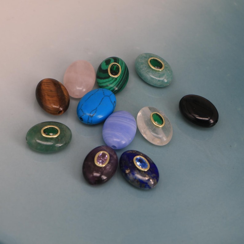 Puede incluir: Una colecci&oacute;n de cuentas de piedras preciosas de forma ovalada en varios colores, incluyendo verde, azul, rosa y marr&oacute;n. Algunas cuentas tienen un anillo dorado alrededor de una piedra m&aacute;s peque&ntilde;a. Las cuentas est&aacute;n dispuestas sobre una superficie azul claro.