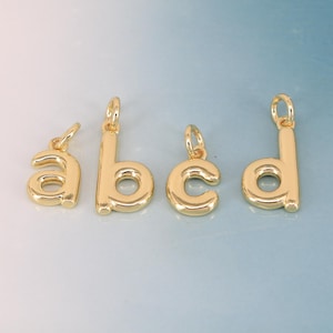Puede incluir: Cuatro colgantes dorados con letras minúsculas: a, b, c y d. Cada colgante tiene un pequeño lazo en la parte superior para sujetarlo a un collar o pulsera. Las letras están en una fuente sencilla y redondeada.