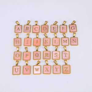 Könnte beinhalten: Eine Sammlung goldfarbener Initialen-Anhänger. Jeder Anhänger hat einen quadratischen, rosa Perlmutt-Inlay mit einem goldenen Buchstaben. Die Anhänger sind in Reihen angeordnet und zeigen das gesamte Alphabet.