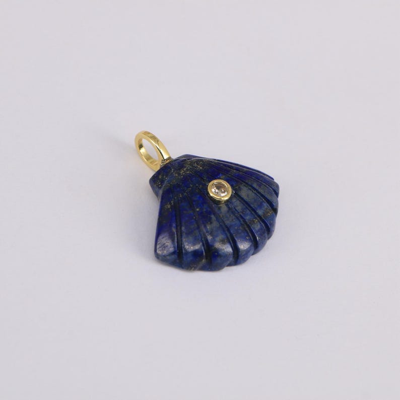 May include: A dark blue lapis lazuli seashell pendant with a gold bail and a small, round, clear stone. The pendant has carved lines and a polished finish, set against a white background.