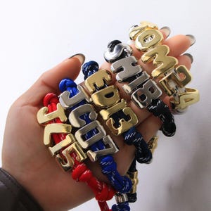 以下が含まれることがあります： 金と銀の金属文字チャームが付いた、パーソナライズされたブレスレットのコレクション。各ブレスレットには、赤、青、黒、白のカラフルなロープバンドが付いています。文字はさまざまな名前と数字を綴っています。