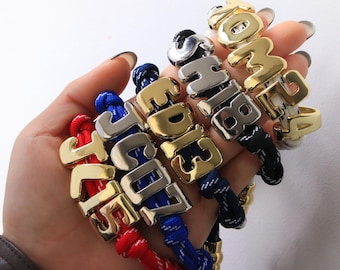 Pulsera anudada con dije de letra y número en relieve, color oro y plata (1 unidad). Joyería personalizada.