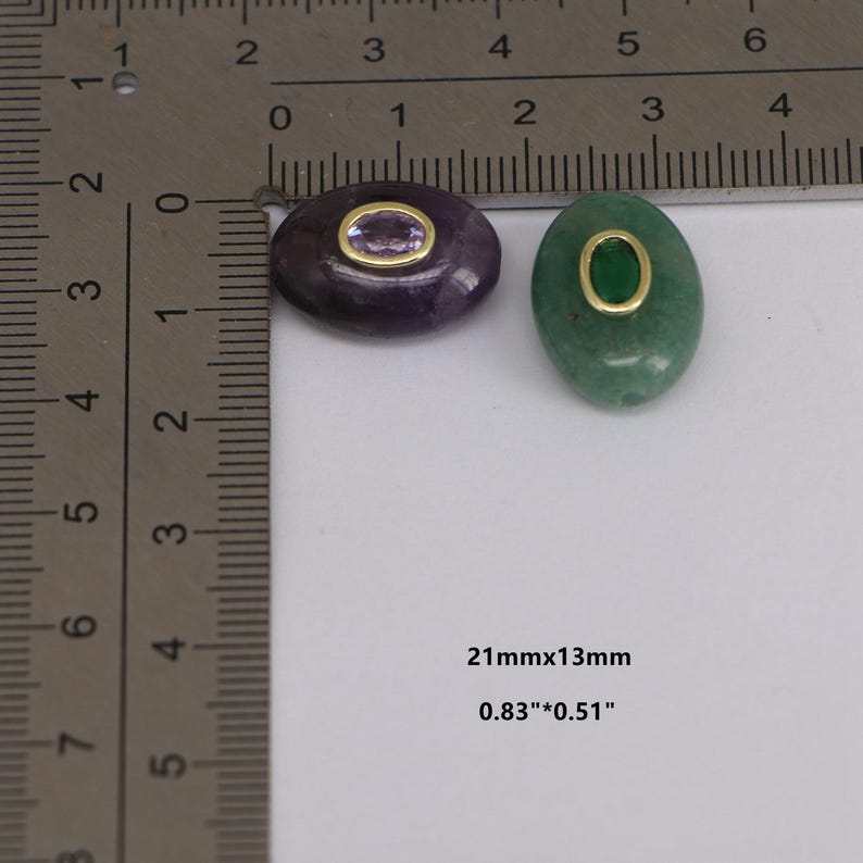 Puede incluir: Dos colgantes ovalados de piedras preciosas. Uno es morado con un centro morado claro, el otro es verde con un centro verde. Ambos tienen bordes de metal dorado. Miden 21mm x 13mm.
