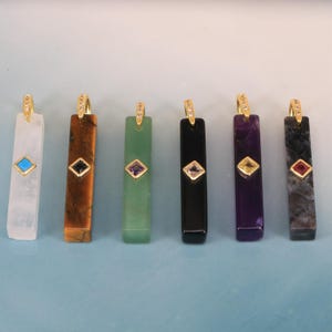 Puede incluir: Seis colgantes rectangulares de piedra en blanco, marrón, verde, negro, morado y gris, cada uno con una anilla dorada con pequeñas piedras claras y una gema cuadrada incrustada en un marco dorado en la parte frontal.