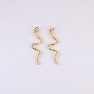 Peut inclure: Deux boucles d'oreilles dorées en forme de serpent. Chaque boucle d'oreille présente un design ondulé et une petite boucle en haut pour l'accrochage. Les boucles d'oreilles sont présentées sur un fond blanc.
