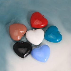 Puede incluir: Una colección de colgantes en forma de corazón en varios colores: rojo, blanco, azul, negro, azul claro y marrón. Cada corazón tiene un pequeño lazo en la parte superior para colgar. Los colgantes son brillantes y lisos.