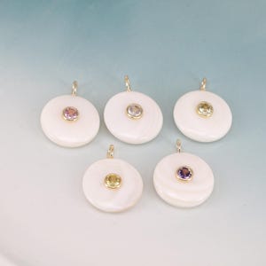 May include: Five circular white shell pendants, each featuring a single bezel-set gemstone in the center. The stones include shades of pink, clear, yellow, and purple. Each pendant has a small gold-toned metal loop at the top for attachment.
