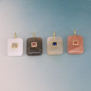 Könnte beinhalten: Vier rechteckige Anhänger mit goldfarbenen Ösen. Jeder Anhänger hat einen andersfarbigen Stein in einem goldfarbenen quadratischen Rahmen. Die Steine sind weiß, rosa, blau und pfirsichfarben. Die Anhänger werden vor einem hellblauen Hintergrund präsentiert.