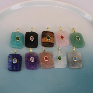 Puede incluir: Una colección de diez colgantes de piedras preciosas en varios colores, incluyendo turquesa, negro, ojo de tigre, rosa y verde. Cada colgante rectangular presenta una argolla dorada y una pequeña gema central. Los colgantes se exhiben sobre una superficie azul claro.