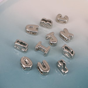 May include: A collection of shiny, silver alphabet beads, including the letters A, B, C, H, I, J, K, O, U, and a stylized letter. The beads are scattered on a light blue surface, perfect for jewelry making or crafting.