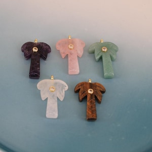 May include: Five gemstone palm tree pendants in shades of purple, pink, green, white, and brown. Each pendant features a gold-colored bail and a small, clear gemstone accent in the center. The pendants are arranged on a light blue surface.