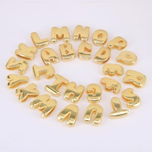 Könnte beinhalten: Eine Sammlung goldfarbener Buchstabenanhänger. Jeder Buchstabe ist in einer abgerundeten, blasenartigen Schriftart gehalten. Die Buchstaben sind in einem kreisförmigen Muster vor einem weißen Hintergrund angeordnet. Diese Anhänger eignen sich für die Schmuckherstellung und das Basteln.