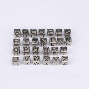 Könnte beinhalten: Silberfarbene Buchstabenperlen, jede würfelförmig mit einem einzelnen Buchstaben auf jeder Seite graviert. Die Buchstaben A bis Z sind abgebildet, perfekt für die Schmuckherstellung oder Bastelprojekte.