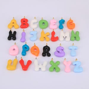 Pode incluir: Uma coleção de pingentes de letras do alfabeto coloridas em estilo bolha. Cada letra tem uma cor diferente, incluindo amarelo, vermelho, branco, verde, rosa, azul, laranja, preto, roxo e azul claro. Os pingentes têm uma argola dourada na parte superior.