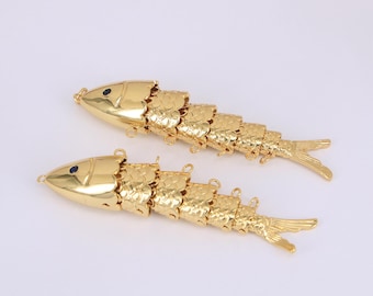 May include: Two gold-colored fish pendants. Each fish has a segmented body, a small eye, and a fin. The pendants are made of metal and have a shiny finish. The fish are lying on a white surface.