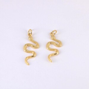 May include: Two gold-colored snake charms with a textured surface. Each charm has a loop at the top for attaching to a necklace or bracelet. The snakes are in a curved, slithering pose, set against a white background.