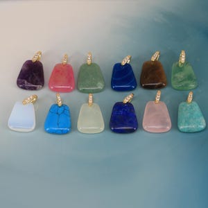 May include: A collection of twelve trapezoidal gemstone pendants arranged in two rows. Each pendant features a polished, smooth stone in various colours including purple, pink, green, blue, brown, white, and turquoise, topped with a gold-tone bail.