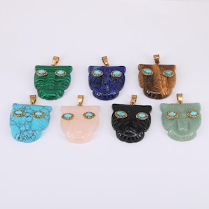 Puede incluir: Colección de colgantes de piedras preciosas tallados con forma de cara de gato estilizada. Cada colgante presenta ojos de piedra de color turquesa y una fianza dorada. Los colgantes son de varios colores, incluyendo verde, azul, rosa y negro.
