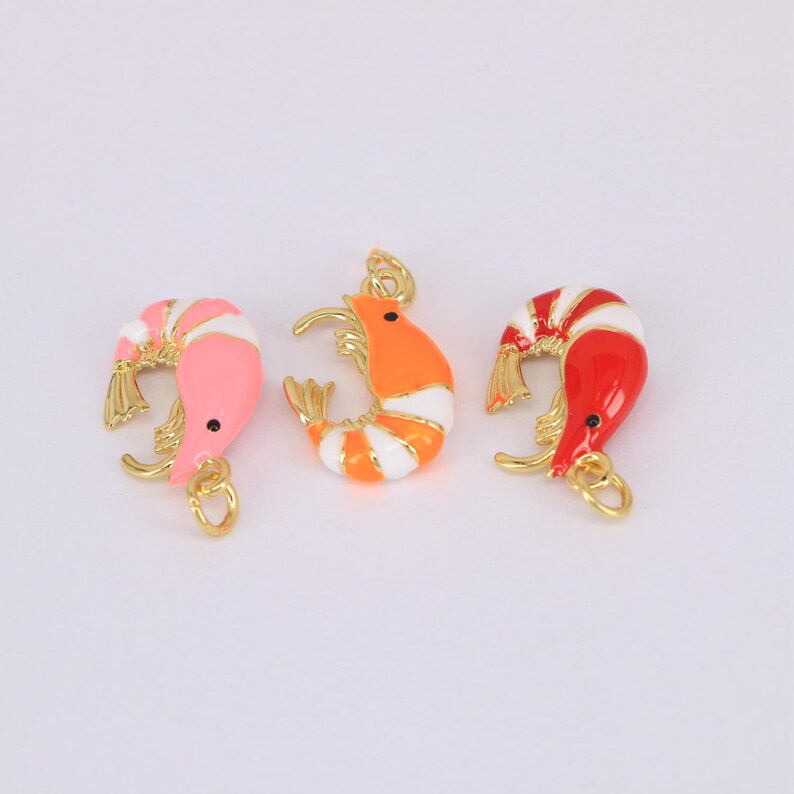 May include: Three colorful shrimp charms in pink, orange, and red with gold-colored accents. Each charm has a small black eye and a gold loop for attaching to jewelry or other crafts. The shrimp have white stripes.