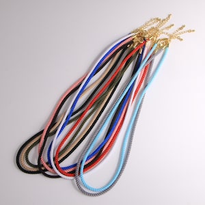 Op de afbeelding: Een verzameling kleurrijke touwkettingen in verschillende tinten, waaronder zwart, roze, wit, blauw, rood en grijs. Elke ketting heeft een goudkleurige sluiting. De kettingen liggen op een witte ondergrond.