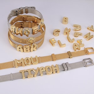 Könnte beinhalten: Gold- und silberfarbene Mesh-Armbänder mit goldenen Buchstaben-Anhängern, die Namen buchstabieren. Einzelne goldene Buchstaben sind in der Nähe verstreut. Die Armbänder haben verstellbare Verschlüsse. Personalisierter Schmuck.