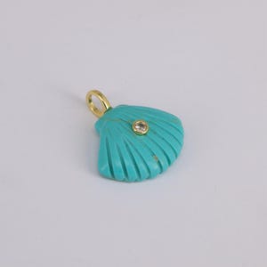 May include: A turquoise seashell pendant with a gold bail and a small, round, clear gemstone. The pendant has carved lines radiating from the center, giving it a textured appearance. The pendant is approximately 1 inch wide.