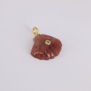 May include: A reddish-brown seashell pendant with a gold-colored loop. The pendant has a small, round, clear stone set in the center. The seashell has a textured surface and is set against a white background.