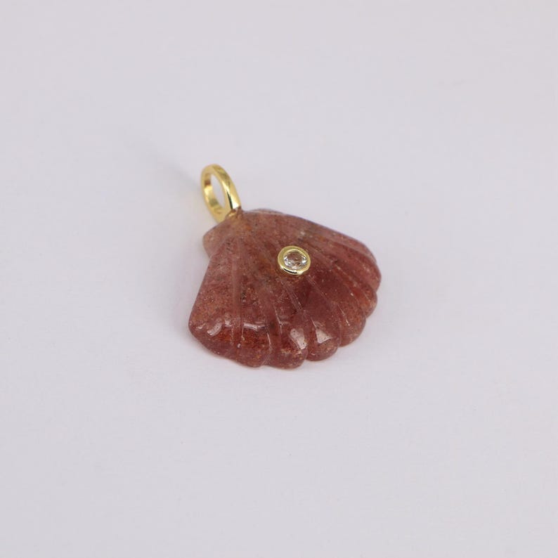 May include: A reddish-brown seashell pendant with a gold-colored loop. The pendant has a small, round, clear stone set in the center. The seashell has a textured surface and is set against a white background.